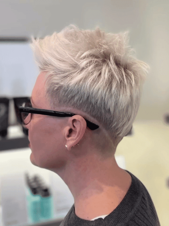 Close-up of a person with a trendy short blonde haircut, wearing glasses, and a dark shirt.