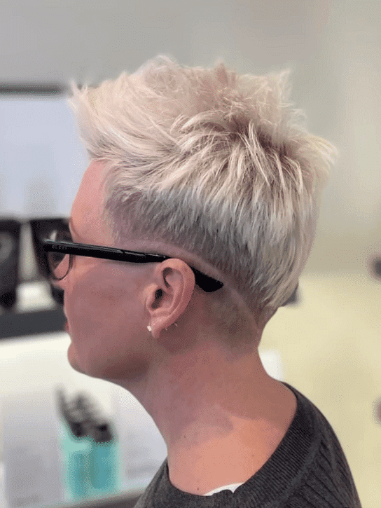 Close-up of a person with a trendy short blonde haircut, wearing glasses, and a dark shirt.