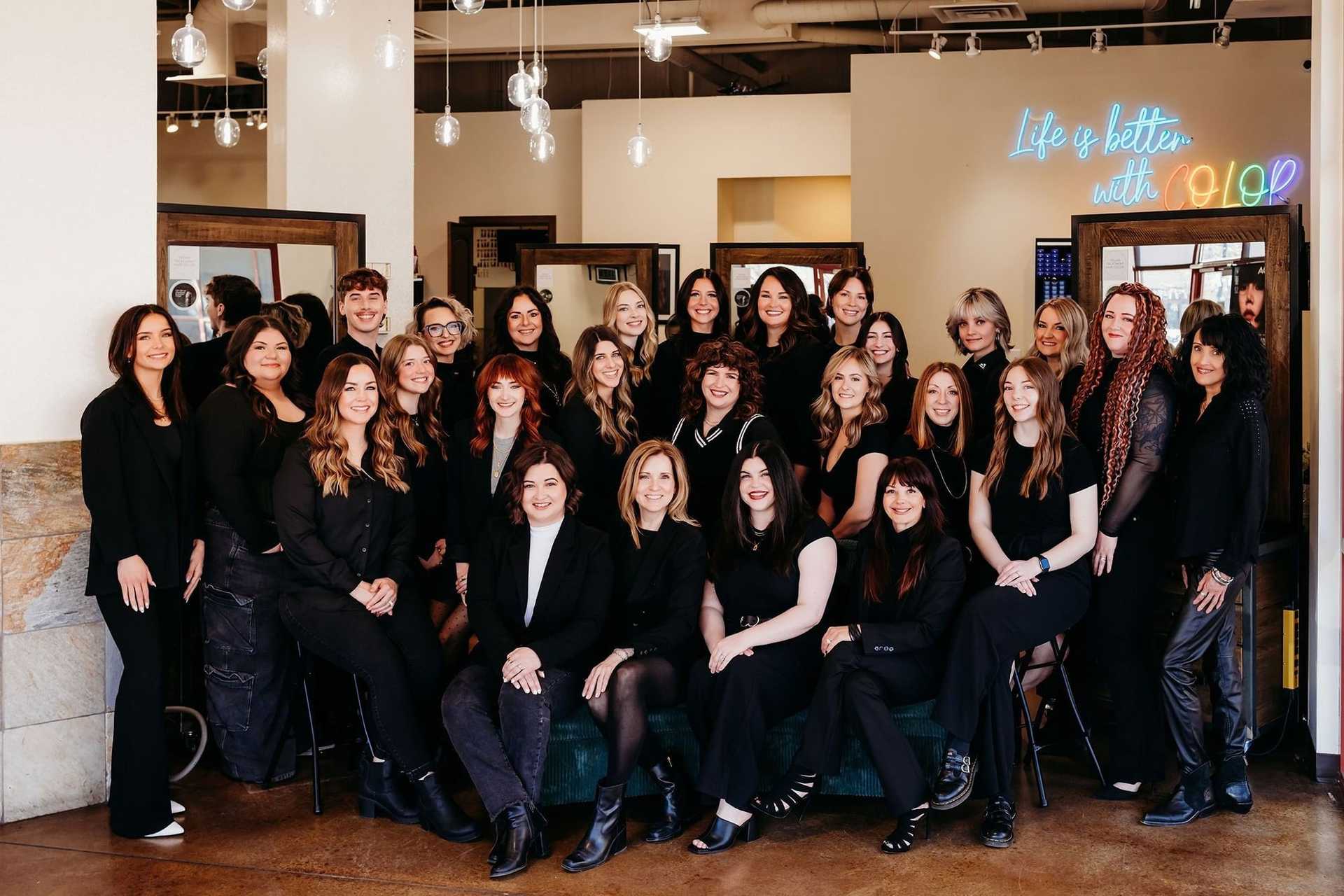 Group of hairstylists in a salon, wearing black outfits, smiling for a team photo.